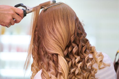 Person curling long, light brown hair with a curling iron in a bright setting.